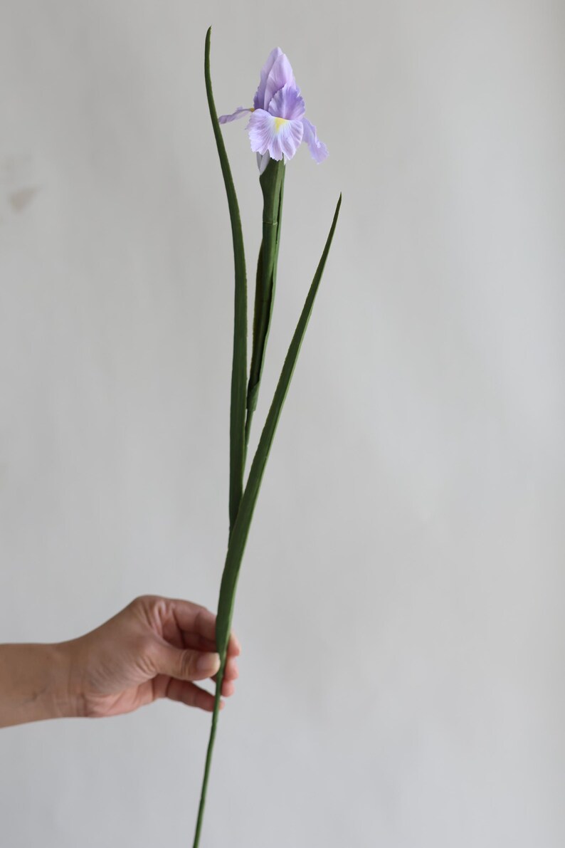 May include: A single artificial purple iris flower with green leaves. The flower is being held in a hand.