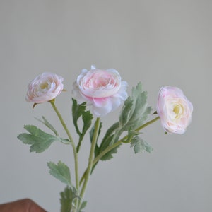 May include: A bouquet of three pink artificial flowers with green leaves. The flowers have a delicate, layered petal design.