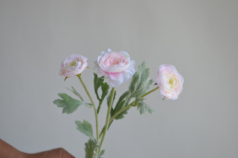 May include: A bouquet of three pink artificial flowers with green leaves. The flowers have a delicate, layered petal design.