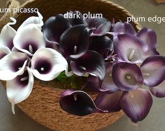 May include: A bouquet of artificial calla lilies in three colors: white with purple accents, dark purple, and a lighter purple with a white edge. The flowers are arranged in a woven basket.