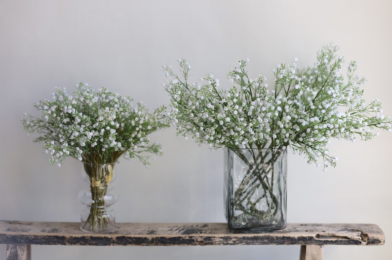 Op de afbeelding: Twee glazen vazen gevuld met witte baby's breath bloemen op een rustiek houten plank.