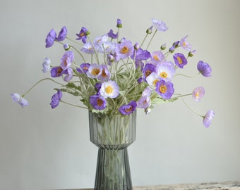 May include: A bouquet of purple and white silk poppy flowers in a clear glass vase with a textured surface.