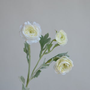 May include: A bouquet of three white artificial ranunculus flowers with green leaves.