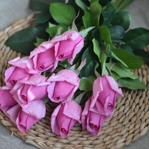 May include: A bouquet of pink silk roses with green leaves arranged on a woven wicker surface.