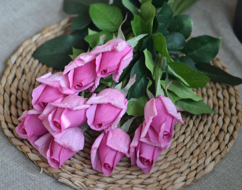 May include: A bouquet of pink silk roses with green leaves arranged on a woven wicker surface.