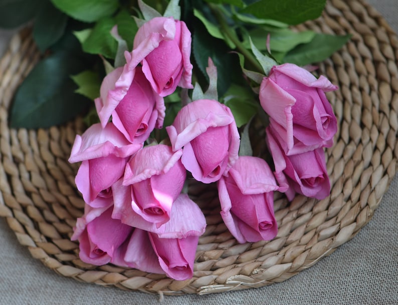 May include: A bouquet of pink artificial roses arranged on a woven wicker surface.