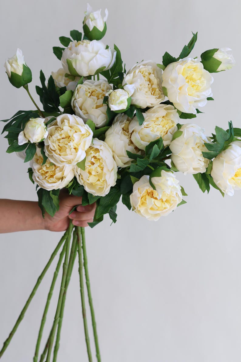 Pu&ograve; includere: Un mazzo di peonie artificiali bianche con foglie e steli verdi.