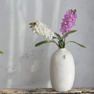 May include: A white ceramic vase with a bulbous shape holds white and purple hyacinth flowers. The vase has a small, round handle on the side. The flowers have green leaves and are set against a neutral background.