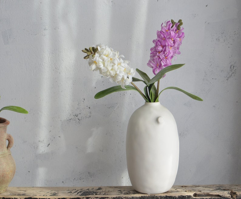 May include: A white ceramic vase with a bulbous shape holds white and purple hyacinth flowers. The vase has a small, round handle on the side. The flowers have green leaves and are set against a neutral background.
