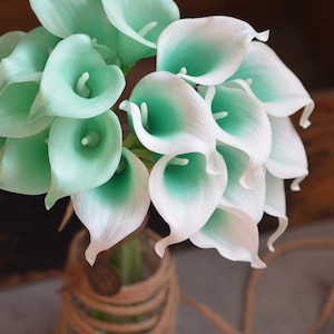 May include: A bouquet of white and light green artificial calla lilies in a clear glass vase with a brown rope wrapped around it.