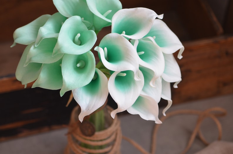 May include: A bouquet of white and light green artificial calla lilies in a clear glass vase with a brown rope wrapped around it.