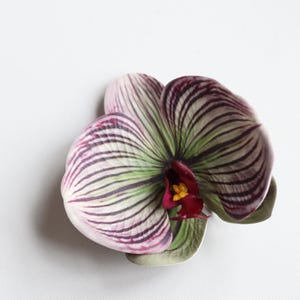 May include: A single artificial orchid flower with white, green, and purple stripes. The flower has a red center with a yellow center.