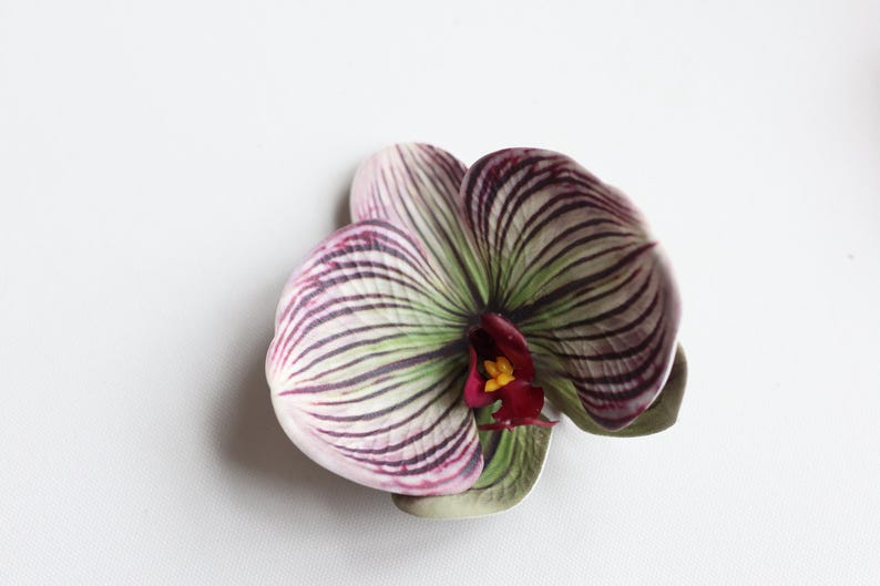 May include: A single artificial orchid flower with white, green, and purple stripes. The flower has a red center with a yellow center.
