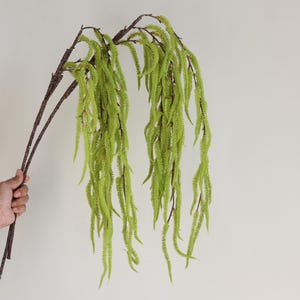 Puede incluir: Una planta artificial decorativa con largos mechones texturizados de color verde brillante en cascada. La planta tiene tallos y ramas de color marrón oscuro. La planta se sostiene sobre un fondo blanco liso.