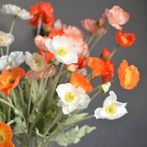 Puede incluir: Un ramo de amapolas artificiales en tonos blancos, naranjas y rojos. Las flores tienen p&eacute;talos delicados y est&aacute;n dispuestas en un jarr&oacute;n.