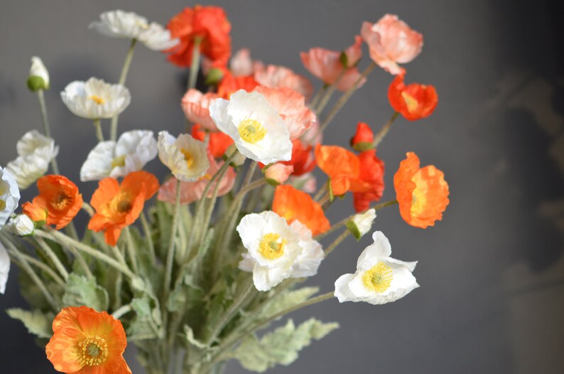 Puede incluir: Un ramo de amapolas artificiales en tonos blancos, naranjas y rojos. Las flores tienen p&eacute;talos delicados y est&aacute;n dispuestas en un jarr&oacute;n.