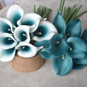 May include: Two bouquets of artificial calla lilies. One bouquet features white petals with teal centers, and the other is solid teal. The flowers are arranged with green stems, and one bouquet is on a burlap-wrapped base.