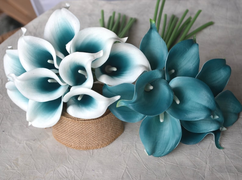 May include: Two bouquets of artificial calla lilies. One bouquet features white petals with teal centers, and the other is solid teal. The flowers are arranged with green stems, and one bouquet is on a burlap-wrapped base.
