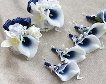 Puede incluir: Un conjunto de siete boutonnières de flor de lirio calla azul marino y blanco con detalles de perlas y adornos de estrellas de mar plateadas. Las boutonnières están dispuestas sobre un fondo de tela beige.