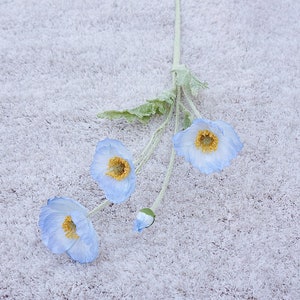 Puede incluir: Un ramo de tres flores de amapola artificiales de color azul claro con centros amarillos. Las flores est&aacute;n dispuestas sobre una superficie blanca y peluda.