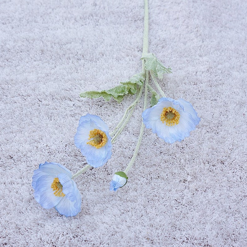 Puede incluir: Un ramo de tres flores de amapola artificiales de color azul claro con centros amarillos. Las flores est&aacute;n dispuestas sobre una superficie blanca y peluda.