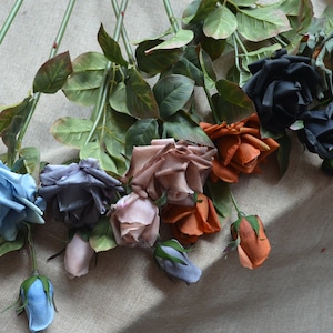Puede incluir: Surtido de rosas de seda artificiales en varios colores, incluyendo azul, gris, rosa, naranja y negro. Las rosas están dispuestas sobre una superficie de tela beige.
