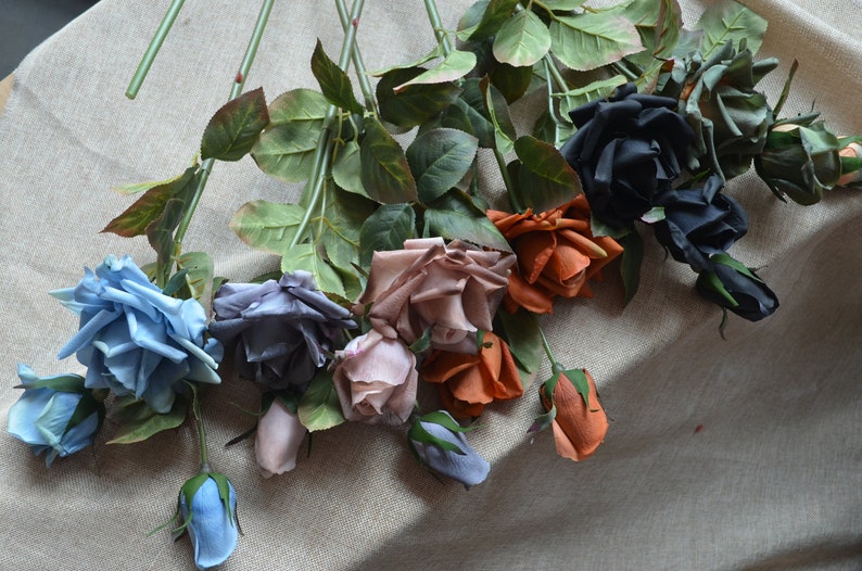 May include: Assortment of artificial silk roses in various colors, including blue, gray, pink, orange, and black. The roses are arranged on a beige fabric surface.