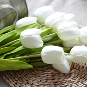 May include: A bouquet of white artificial tulips with green stems and leaves. The tulips are arranged on a woven wicker surface.