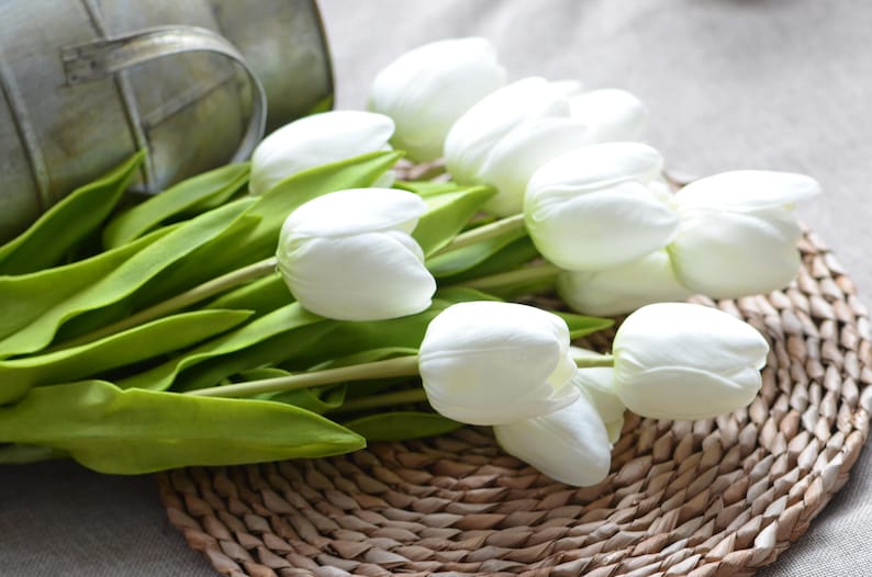 May include: A bouquet of white artificial tulips with green stems and leaves. The tulips are arranged on a woven wicker surface.