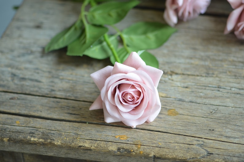 May include: A single pink rose with a green stem and leaves on a wooden surface.