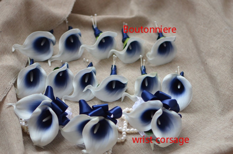 May include: A set of 12 white and navy blue calla lily boutonnieres with navy blue ribbon accents. The boutonnieres are arranged on a beige fabric background. The text "Boutonniere" is written in red on the top of the image. The text "wrist corsage" is written in red on the bottom of the image.