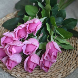 May include: A bouquet of pink artificial roses with green leaves, arranged on a woven wicker surface.