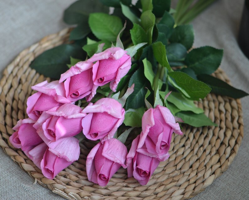 May include: A bouquet of pink artificial roses with green leaves, arranged on a woven wicker surface.
