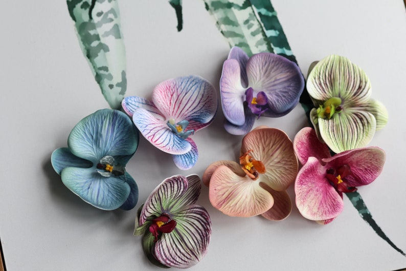 May include: Seven artificial orchid flowers in various colors, including blue, pink, purple, green, and peach. The flowers are arranged on a white surface.