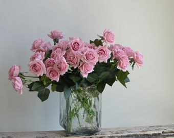 May include: A bouquet of pink silk roses arranged in a clear glass vase with a textured surface. The vase is sitting on a weathered wooden surface.