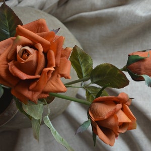 May include: Three artificial orange roses with green leaves. The roses are in various stages of bloom, with one fully open, one partially open, and one still in bud.