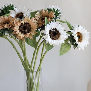 Op de afbeelding: Een heldere glazen vaas bevat een boeket kunstmatige zonnebloemen. De zonnebloemen hebben witte, bruine en bruine bloemblaadjes met donkerbruine centra. Groene bladeren en stelen strekken zich uit van de bloemen. De achtergrond is licht van kleur.