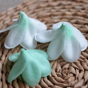 May include: Three fabric flower shapes, two white with green edges and one green with white edges, are arranged on a woven brown basket surface.
