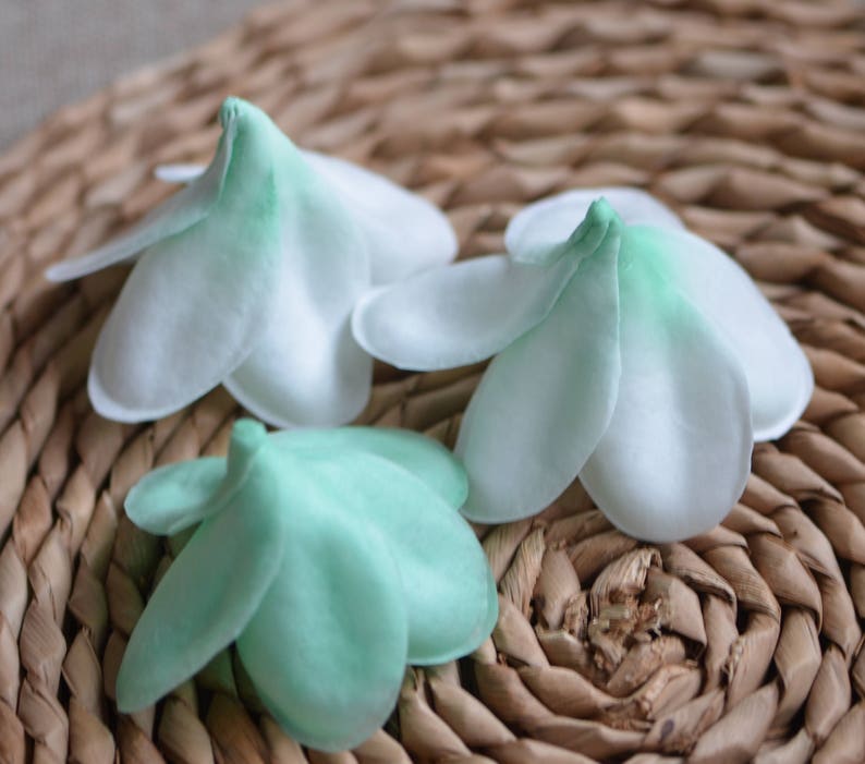 May include: Three fabric flower shapes, two white with green edges and one green with white edges, are arranged on a woven brown basket surface.