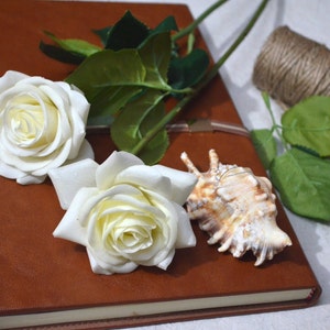 May include: A brown leather journal with two white roses and a seashell on top. The journal is closed and has a white ribbon tied around it.