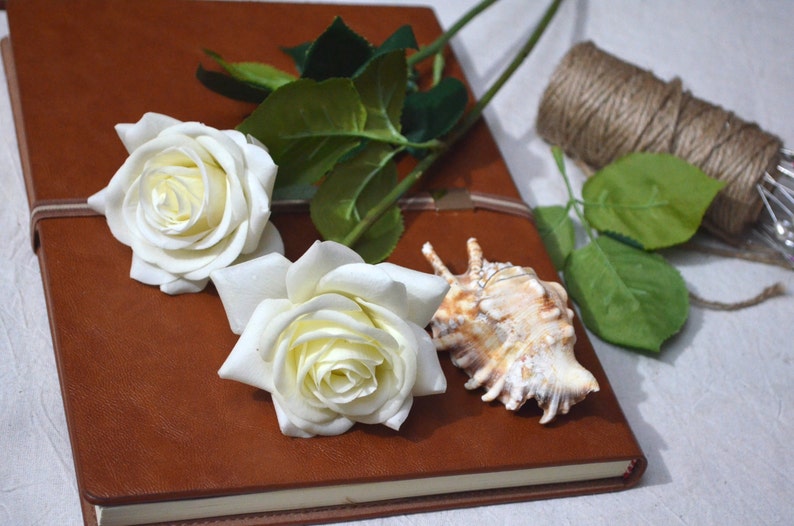 May include: A brown leather journal with two white roses and a seashell on top. The journal is closed and has a white ribbon tied around it.