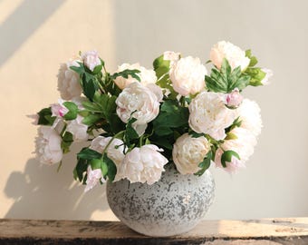 Pode incluir: Um buquê de peônias brancas e rosadas em um vaso de cerâmica cinza manchado. As flores são dispostas em um estilo solto e natural.