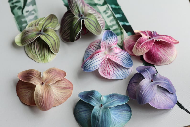 May include: Assortment of seven artificial orchid flower heads in various colors, including green, pink, blue, and purple. The flowers are made of fabric and have a realistic appearance.