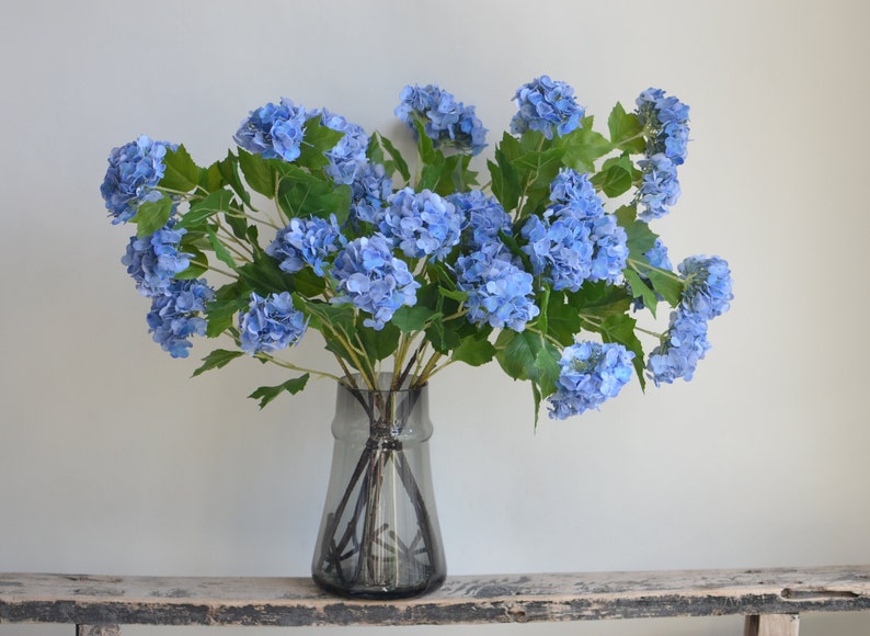 May include: A bouquet of blue hydrangeas in a clear glass vase with a textured surface. The vase is sitting on a wooden surface.