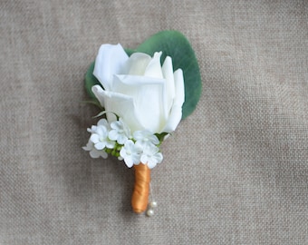Puede incluir: Un boutonniere de rosa blanca con pequeñas flores blancas y hojas verdes, atado con una cinta dorada y detalles de perlas.