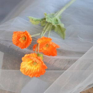 Puede incluir: Un ramo de cuatro flores de amapola de seda naranja con hojas verdes sobre un fondo de tul blanco.