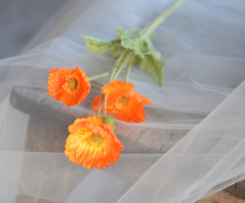 Puede incluir: Un ramo de cuatro flores de amapola de seda naranja con hojas verdes sobre un fondo de tul blanco.