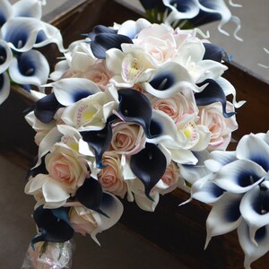 May include: A cascading bridal bouquet featuring white and black calla lilies, blush pink roses, and white orchids. The bouquet is tied with a white ribbon.