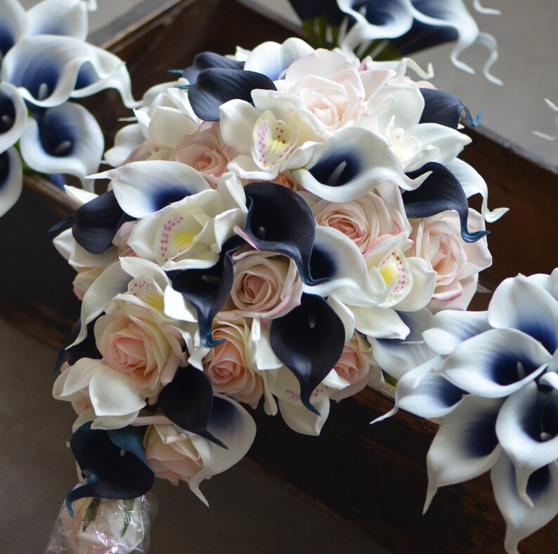 May include: A cascading bridal bouquet featuring white and black calla lilies, blush pink roses, and white orchids. The bouquet is tied with a white ribbon.
