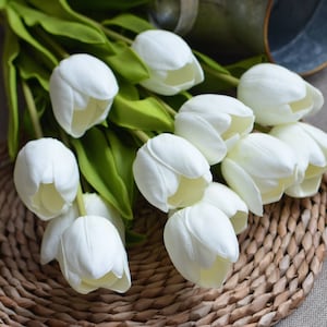 May include: A bouquet of white artificial tulips arranged on a woven wicker placemat. The tulips are made of a soft, realistic material and have a natural, elegant look.